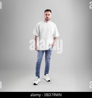 Mockup di t-shirt oversize bianca su uomo con barba in jeans blu, a mano in tasca, a tutta lunghezza, vista frontale, isolata sullo sfondo studio. Fotog. Prodotto Foto Stock
