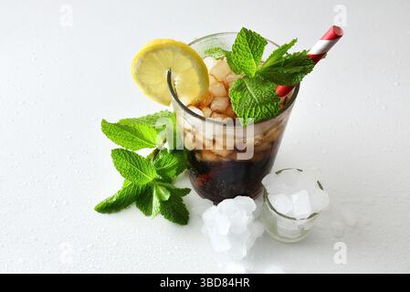 Bevanda rinfrescante con ghiaccio in una tazza di vetro decorata con una paglia e una fetta di frutta su un tavolo bianco con ghiaccio e foglie di menta. Vista rialzata. Foto Stock