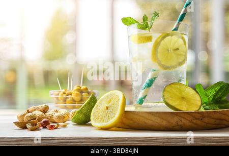Rinfrescante bevanda al limone gassato su un tavolo di legno con spuntini come arachidi e olive decorate con foglie di menta e lime e fette di limone su una t Foto Stock