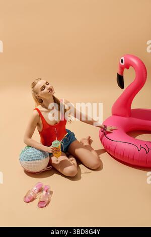 Una giovane donna con la vivace cima rossa si rilassa sul pavimento con un drink e un divertente fenicottero gonfiabile nelle vicinanze. Foto Stock