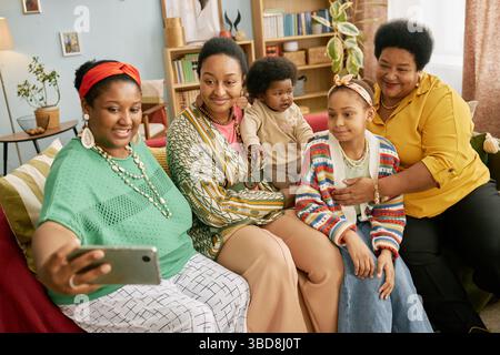 Felice famiglia afroamericana multigenerazionale di bambini e adulti seduti sul divano, sorridendo e scattando selfie con lo smartphone in salotto Foto Stock