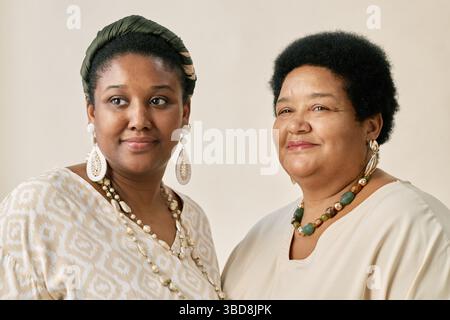 Ritratto di due donne afroamericane che si mettono insieme indossando abiti e accessori tradizionali mentre sorridono per il ritratto in studio contro il neutro Foto Stock