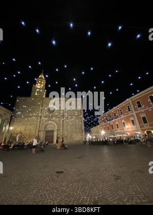 Illuminazione notturna che mette in risalto la facciata ornata della Cattedrale di Atri e il campanile, gettando un caldo bagliore sulla storica Piazza Duomo con i visitatori che si fermano Foto Stock