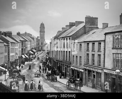 Una fotografia dei primi anni del XX secolo di negozi e acquirenti in Church Street a Enniskillen, la più grande città della contea di Fermanagh, Irlanda del Nord. In cima si può vedere la torre del municipio, progettata da William Scott e completata nel 1901. Foto Stock