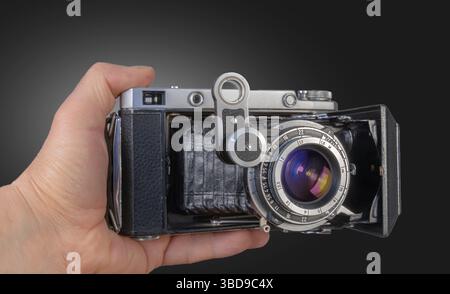Una mano tiene in mano una fotocamera vintage, che mette in mostra il suo complesso obiettivo e il design del corpo in un ambiente ben illuminato che ne migliora le caratteristiche. Foto Stock
