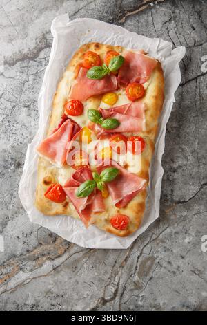 pinsa italiana fatta in casa con pomodori ciliegini, prosciutto, mozzarella e basilico su carta pergamena in tavola. Vista dall'alto verticale Foto Stock