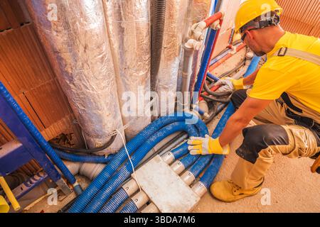 Un operaio edile con una camicia gialla e un casco sta installando con cura tubi idraulici in un sito di ristrutturazione. Vari tubi blu e grigi si intrecciano Foto Stock
