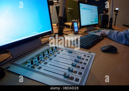 Una sala di controllo affollata dotata di diversi monitor per computer, una console di mixaggio audio e un'emittente che regola le impostazioni durante una sessione radio dal vivo. BR Foto Stock