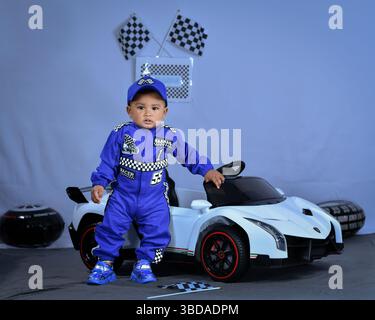 Un bambino vestito da pilota di auto da corsa siede su un'auto sportiva giocattolo per festeggiare il suo primo compleanno, con decorazioni a tema racing. Foto Stock