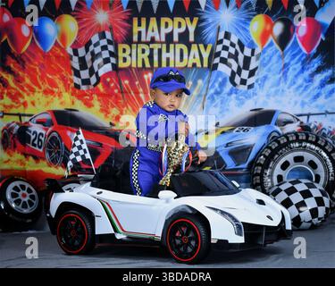 Il bambino festeggia il suo primo compleanno vestito da pilota di auto da corsa, reggendo un trofeo mentre è seduto in un'auto da corsa giocattolo. Foto Stock