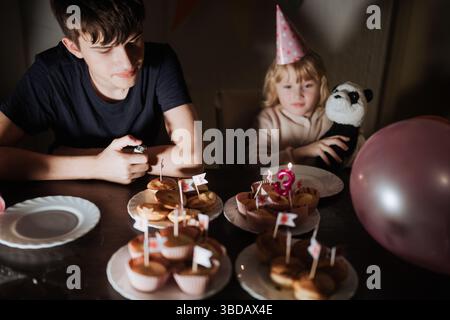 Due fratelli e sorelle celebrano il compleanno con cupcake decorati sul tavolo. Un fratello tiene in mano un panda di peluche mentre l'altro guarda in anticipo Foto Stock