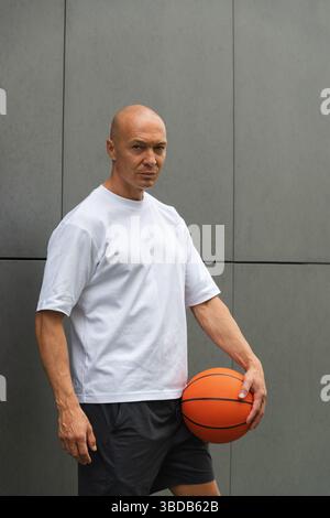 Ritratto di un affascinante giocatore di basket sportivo con t-shirt bianca che tiene la palla e lo guarda vicino al muro grigio. Formato verticale. Foto Stock