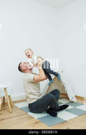 In un accogliente soggiorno, un padre gioioso solleva il suo bambino sorridente, creando un'esperienza giocosa e divertente piena di momenti familiari e di persone Foto Stock