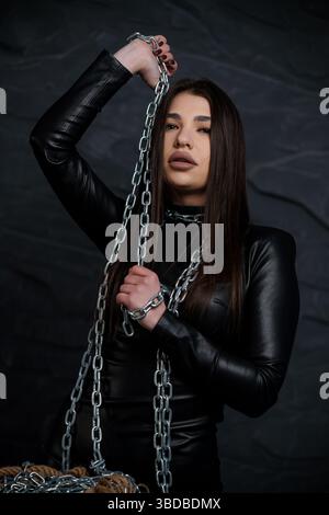 Un suggestivo ritratto di una giovane donna dalla pelle chiara con lunghi capelli scuri, rivestita in pelle nera, che regge una pesante catena argentata contro una pelle scura Foto Stock