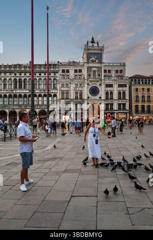 Un turista che dà da mangiare ai piccioni in Piazza San Marco, con la Torre dell'Orologio sullo sfondo, in estate al tramonto, Venezia, Veneto, Italia Foto Stock