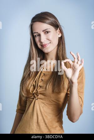 La giovane bella donna mostra il segno OK sullo sfondo blu Foto Stock