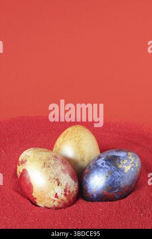 3 uova di Pasqua colorate, parzialmente dorate, su ghiaia decorativa rossa Foto Stock