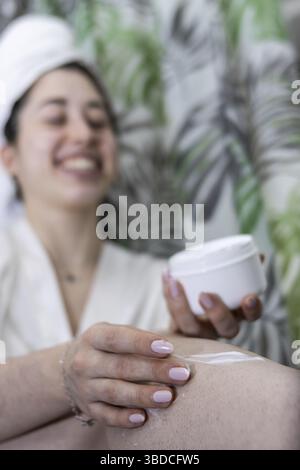 Ritratto verticale di una donna felice che applica crema per il corpo sulle gambe dopo aver fatto una doccia. Messa a fuoco selettiva sul primo piano Foto Stock