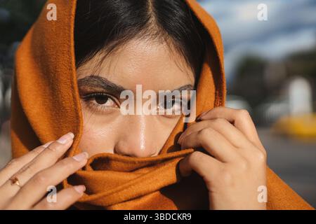 Giovane donna indigena che copre il viso con scialle arancione su sfondo sfocato. moda ritrattistica Foto Stock