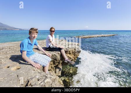 Madre e figlio come turisti che si siedono su roccia al blu del mare godendo di vacanza Foto Stock