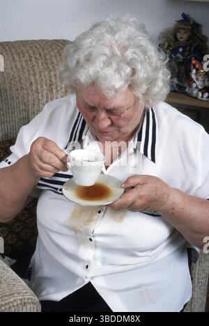 Donna anziana, poltrona, caffè versato, tazza di tè, fuoriuscita, agitare, stringere le mani, tremare, parkinson, morbo di parkinson, sindrome del tremore, tremore par Foto Stock