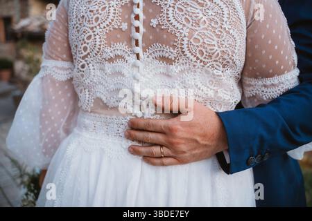 Primo piano di una sposa con un abito da sposa in pizzo e uno sposo con un abito blu, che mette in risalto il loro abbraccio. La mano dello sposo poggia affettuosamente sulla sposa Foto Stock