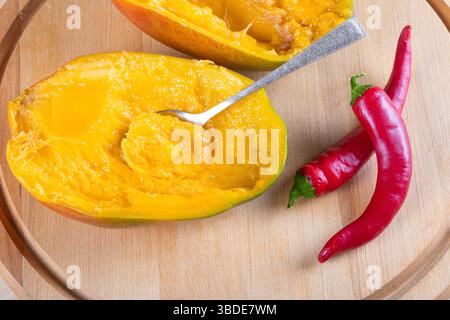 Vista dall'alto di due metà di mango, due peperoncini rossi caldi e cucchiaio su sfondo di legno Foto Stock