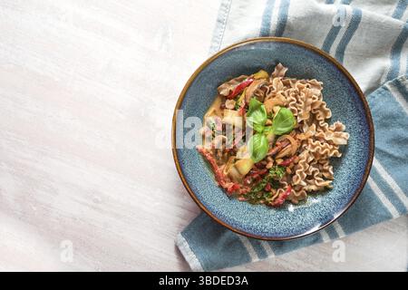 Piatto salutare di pasta integrale al farro con verdure in salsa di pomodoro e basilico in un piatto blu su un tavolo di legno bianco, vista dall'alto Foto Stock