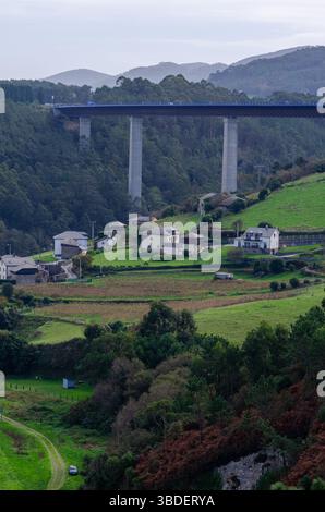 Uno spettacolare ponte autostradale sopraelevato si estende sopra un tranquillo villaggio rurale con lussureggianti campi verdi e colline boscose circostanti, evocando tranquillità Foto Stock