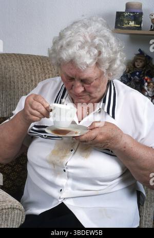 Donna anziana, poltrona, caffè versato, tazza di tè, fuoriuscita, agitare, stringere le mani, tremare, parkinson, morbo di parkinson, sindrome del tremore, tremore par Foto Stock