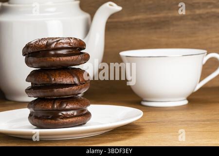 Vista ravvicinata di una deliziosa torta al cioccolato fatta in casa in un piatto di ceramica bianca e una tazza di tè caldo con bollitore per tè bianco su un tavolo di legno Foto Stock