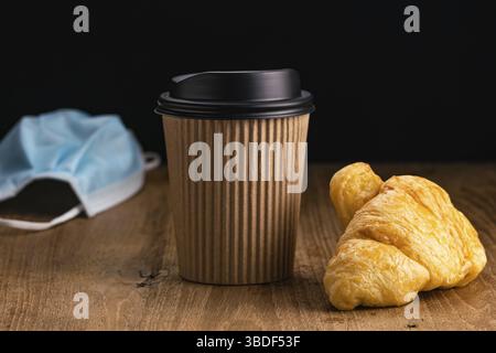 Vista laterale di delizioso croissant con caffè caldo da portare a casa in tazza di carta monouso e maschera medica viso per protezione Covid 19 su tavolo di legno su bl Foto Stock