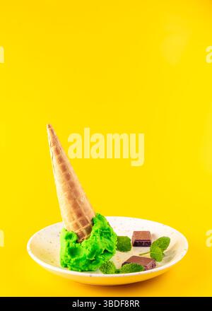 Un primo piano di un gelato verde che si scioglie in un cono, con foglie di menta fresca e pezzi di cioccolato sul piatto accanto Foto Stock