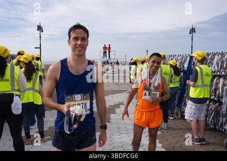 Due corridori maschi, uno in blu e uno in un singlet arancione, stanno vicino al traguardo di una gara. I volontari in gilet gialli sono visibili, lungo l'arguzia Foto Stock