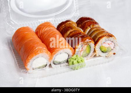Primo piano di sushi rotondi di salmone e anguilla freschi con wasabi in un vassoio di plastica trasparente su una superficie bianca. Sushi, consegna del cibo Foto Stock