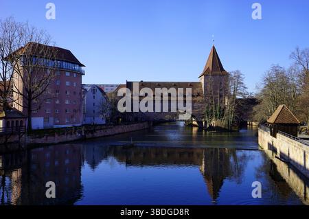 Torri storiche e edifici in legno si ergono lungo un fiume calmo, riflesso nell'acqua sotto un cielo azzurro a Norimberga, in Germania Foto Stock