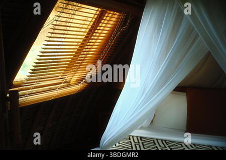 Un comodo letto con tende bianche a baldacchino accanto ad una finestra coperta da persiane di legno Foto Stock