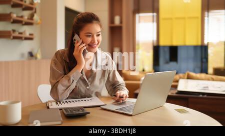 La giovane donna asiatica lavora felicemente a casa utilizzando il laptop e parlando sullo smartphone guarda lo schermo e sorride mentre fa affari e gestisce re Foto Stock