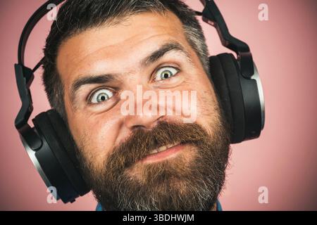 Uomo barbuto divertente che ascolta la musica con le cuffie wireless, ritratti ravvicinati. Bellissimo ragazzo sorridente con barba e baffi che si diverte con le canzoni preferite Foto Stock