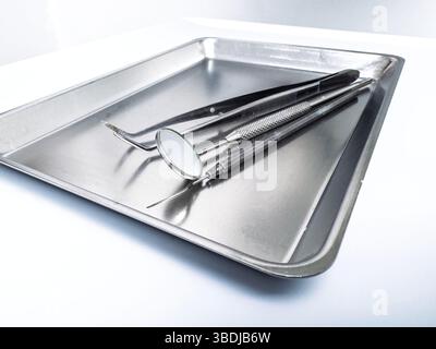 Strumenti odontoiatrici in acciaio inox, tra cui specchio e sonde poggiate su un vassoio in metallo sterile posizionato su una superficie bianca in un ambiente clinico illuminato Foto Stock