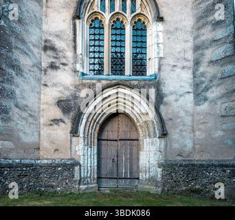 Primo piano di una pesante porta di chiesa ad arco in legno con elementi in ferro, posta sotto una finestra di vetro al piombo all'interno di un muro di pietra temprata che ha avuto Foto Stock