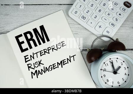 Testo di ERM Enterprise Risk Management su pagina notebook cartacea con sveglia e calcolatrice su sfondo in legno Foto Stock