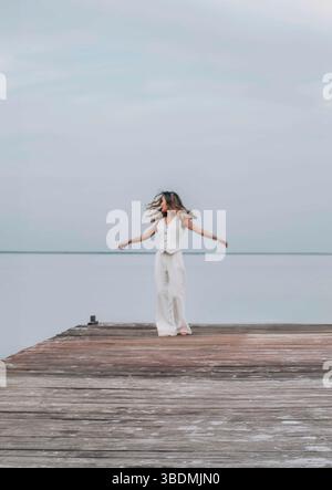 donna che danzava a piedi nudi su un molo di legno, capelli lunghi che scorrevano liberamente sotto un cielo pallido, circondato da acqua calma, espressione gioiosa Foto Stock