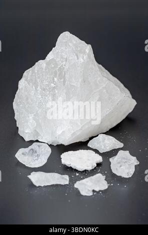 Un grande frammento di cristallo di alluminio bianco è esposto su una superficie scura, circondato da piccoli frammenti di cristallo, che mostrano la sua naturale consistenza e chiarezza Foto Stock