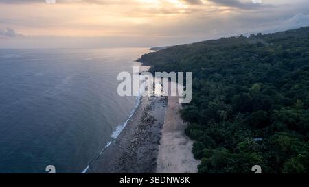Vista aerea di una costa tropicale durante il tramonto, che mostra la lussureggiante foresta, la spiaggia sabbiosa, la barriera corallina e le calme onde dell'oceano. Foto Stock