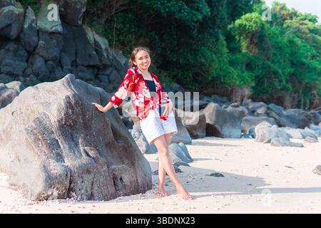 La giovane donna indonesiana sorride mentre si appoggia su una grande roccia in una spiaggia tropicale. Foto Stock