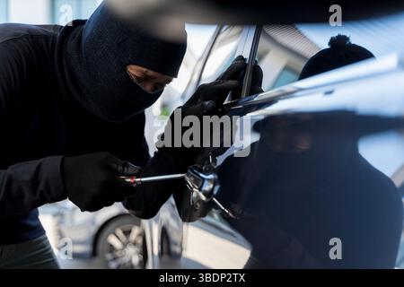 Un ladro mascherato che indossa guanti e utilizza un cacciavite per entrare nella porta di un'auto in pieno giorno. Concetto di furto del veicolo, violazione della sicurezza, pericolo, crimine Foto Stock
