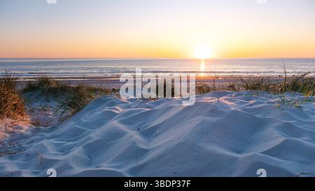 La vista del tramonto proietta vivaci sfumature sulla tranquilla spiaggia di Vejers Strand, Danimarca. Le morbide dune di sabbia adornate di erbe incorniciano la costa, dove le onde si infrangono dolcemente contro la riva. Foto Stock