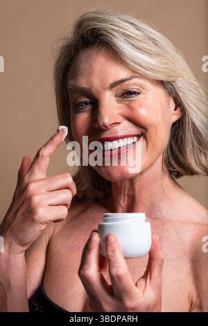 Ritratto di bellezza di una bella donna matura, concetti su cosmetici, cura della pelle e benessere. Donna matura sorridente con capelli biondi corti applicando il viso c Foto Stock