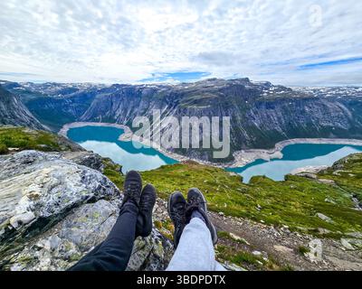 Godendoti la vista mozzafiato da una scogliera aspra, due paia di gambe sporgono dal bordo. Sotto, i laghi turchesi brillano sotto le imponenti vette delle montagne. Escursione Trolltunga Norvegia Foto Stock